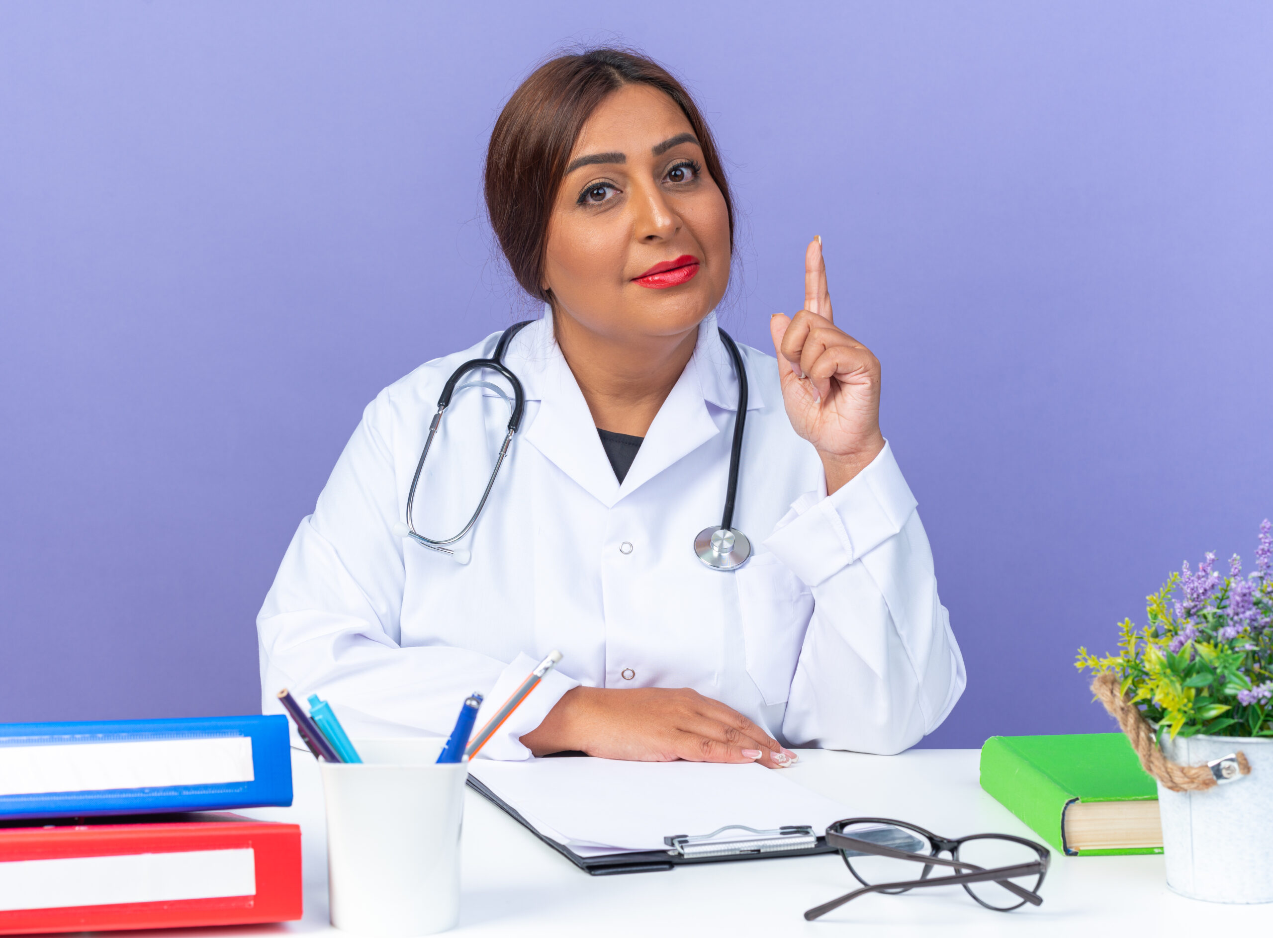 middle age woman doctor in white coat with stethoscope looking at camera with confident expression showing index finger like warning sitting at the table over blue background